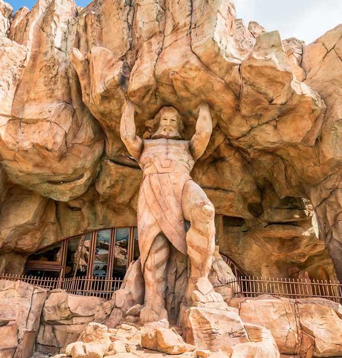 An image of a rock formation on the exterior of Mythos. It is light brown, and has a gate with a half-circle of paneled windows. There is a rock formation of a  man wearing what appears to be a toga, who has long hair, a mustache, and a beard, appearing to be holding up the building's rock formation.