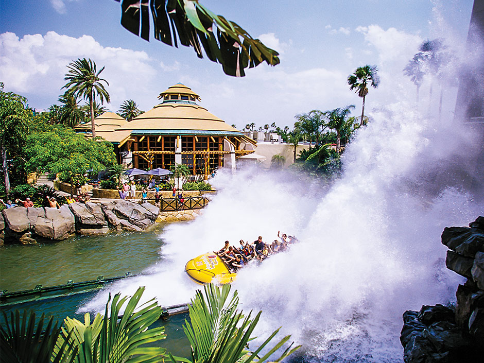 A wide-view of a yellow raft full of people finishing the drop on Jurassic Park River Adventure. Giant splashes of water surround the vehicle, landing in the river on either side of the ride track. Behind the river is the Thunder Falls Terrace building. Several people stand at various rock formations and gates separating the terrace from the ride's lake, waving. Many trees surround the building. The sky is blue and full of white clouds.