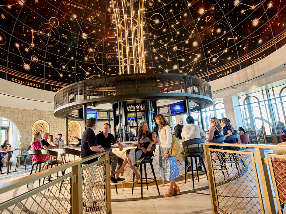 A group of friends stand and chat at the bar in Bar Helios at Helios Grand Hotel
