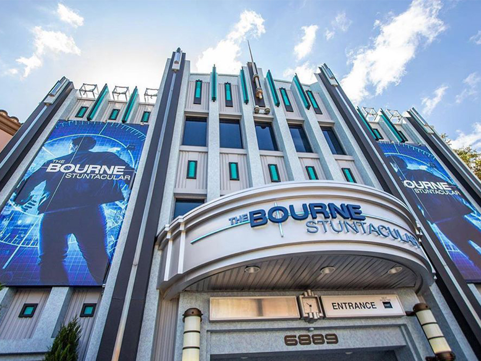 The Bourne Stuntacular building's entrance on a relatively clear day. The building has three connected towers, with the two side towers displaying signs reading "The Bourne Stuntacular" in white in front of a man in a jacket's back. On the signs, the man stands against a blue, gridded background, and has a circular target surrounding and slightly covering him. The silver-colored marquee on the center building reads "The Bourne Stuntacular" with the first and last words in light blue, and the middle word in darker blue and larger font. Under the marquee is an empty sign on the left, a clock in the center, and a sign on the right that reads "Entrance." Under that is brown text reading "6 8 8 9." Surrounding the sign and numbers are two cylindrical lamps. Above the marque are two rows of seemingly five, rectangular windows each, though only one window (and the corner of a second) are visible from the lower row. On all three buildings are thin, turquoise panels with dark borders. A stick with a pointed top sits at the center and top of the center tower.