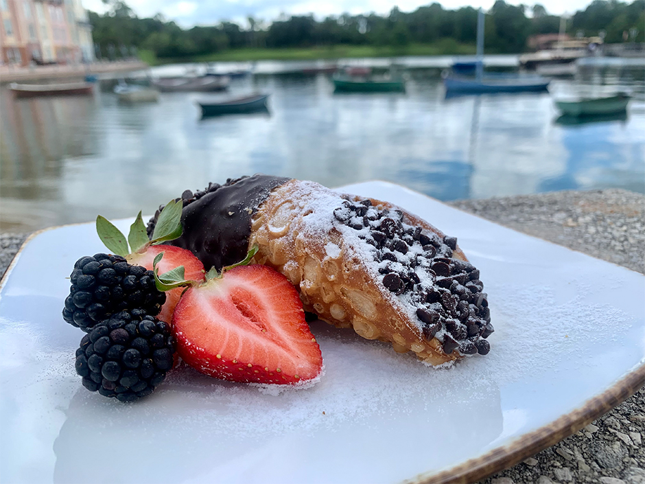 A Chocolate Dipped Cannoli filled with chocolate chips and covered in powdered sugar sitting on a white plate, also covered in powdered sugar. Two halved strawberries and two blackberries sit next to the cannoli. Portofino Harbor is visible in the background.