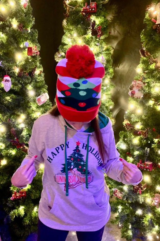 Woman wearing winter hat and holiday sweatshirt with Christmas tree in background