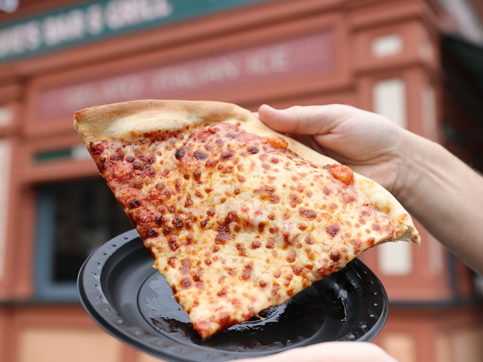 Hand Holding Slice of Cheese Pizza with Louie's restaurant in the background