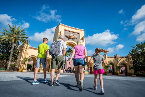 A family walking toward the beige arches at the entrance to Universal Studios Florida. From left to right: a man in a bright green shirt, gray shorts, and sneakers in various shades of blue with a white lining; an older man in a gray shirt, green shorts, and blue sneakers with a white lining, with a gray-ish-blue backpack; a woman wearing a light purple shirt, jeans, gray sneakers, and a white wristband on her left wrist; a girl with pigtails wearing a gray shirt and pink shorts. The outer arches are shorter than the one in the center, which has a gray center and the globe logo with text "Universal Studios" in silver and "Florida" in orange, with a line in between. All of the arches have a ridge-patterned straight line at their tops. On either side of the arches are plants, including palm trees, and there is a brown fence on the arches' right. Pieces of buildings are visible behind the arches. The sky is blue with a couple clouds.
