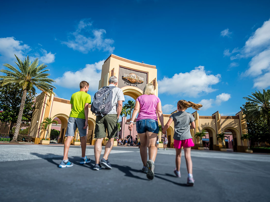 A family walking toward the beige arches at the entrance to Universal Studios Florida. From left to right: a man in a bright green shirt, gray shorts, and sneakers in various shades of blue with a white lining; an older man in a gray shirt, green shorts, and blue sneakers with a white lining, with a gray-ish-blue backpack; a woman wearing a light purple shirt, jeans, gray sneakers, and a white wristband on her left wrist; a girl with pigtails wearing a gray shirt and pink shorts. The outer arches are shorter than the one in the center, which has a gray center and the globe logo with text "Universal Studios" in silver and "Florida" in orange, with a line in between. All of the arches have a ridge-patterned straight line at their tops. On either side of the arches are plants, including palm trees, and there is a brown fence on the arches' right. Pieces of buildings are visible behind the arches. The sky is blue with a couple clouds.