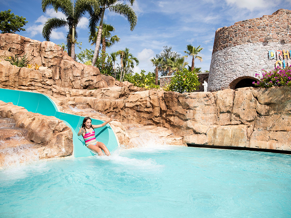 A girl sliding down at the bottom of a turquoise waterslide, into a pool. The girl is wearing a multicolored, striped bathing suit, and her hands are out in front of her in diagonals. She is smiling. The waterslide is surrounded by rock structures on all sides but the front, which goes into the pool, and on one of the rock structures behind it sit a couple plants. Behind the rocks are various trees and bushes. To the slide's left, on top of the rocks, is a stone building with multi-colored text that is cut-off. In front of that is a green bush with red flowers. The sky is blue with several white clouds.