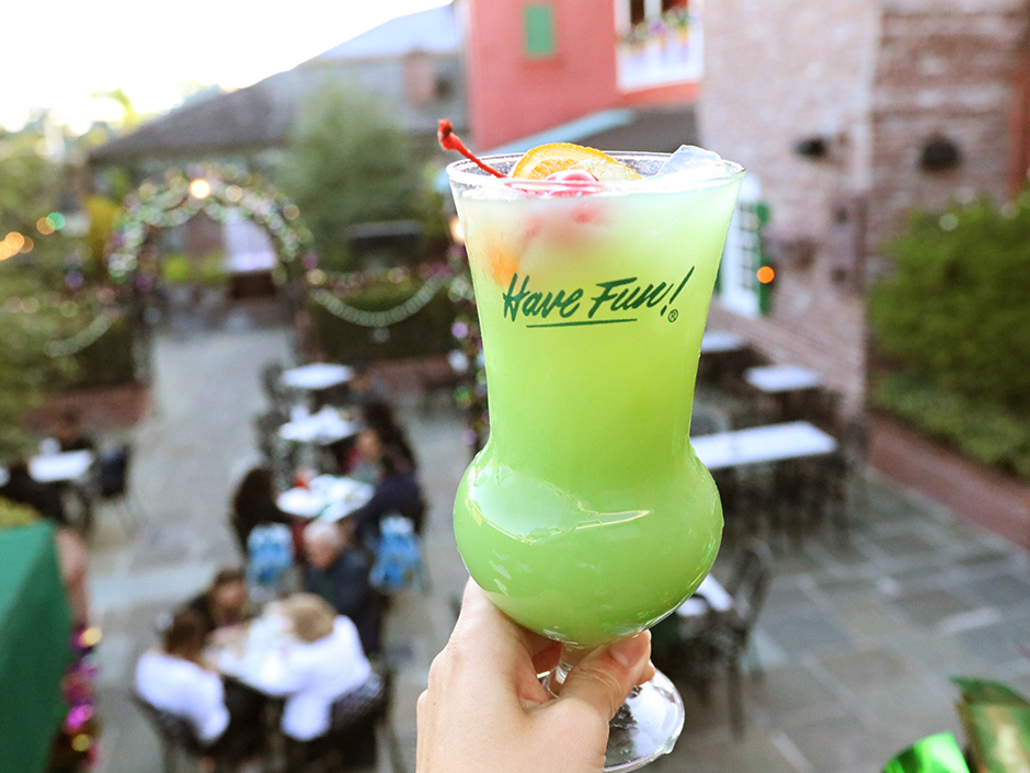 A hand holds up a green drink with ice, an orange, and a cherry, in a glass cup with a circular bottom. The cup reads "Have fun!" with an exclamation point, with the text underlined and in green. The background of people sitting at outdoor tables to the drink's right, and empty tables to the drink's left, seemingly a level below is blurred, as well as the plants, buildings, and sunny sky.