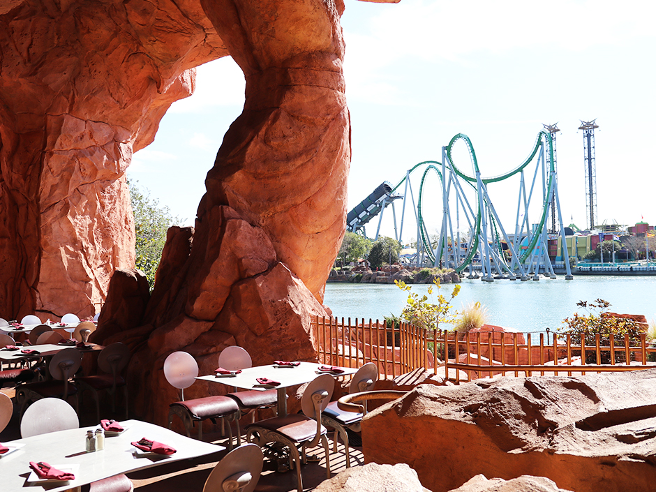 Outdoor tables at Mythos restaurant, overlooking a body of water, on which stands The Incredible Hulk Coaster. The tables visible all have four seats, a plate and folded red napkin at each spot. Various brown rock formations, as well as a fence, line the edge between the seating and the water.