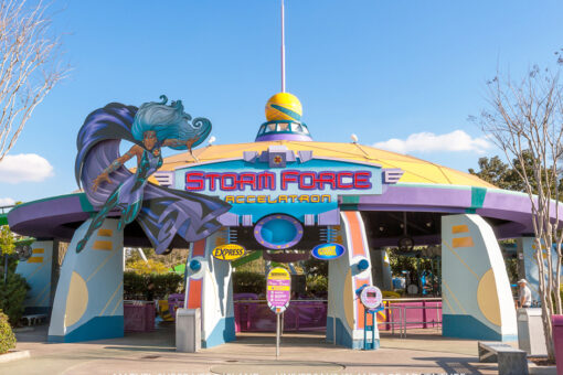 The entrance to Stormforce Accelatron during the daytime. The words "Storm Force" are in red with a blue-ish-purple border, and the word "Accelatron" is in yellow, with a blue gradient background behind bot. There is a blue circle with a purple border with various small, orange shapes under the sign, between a circular, yellow sign with blue text "Express" on the let and a blue sign with yellow text "attraction entrance" on the right. There is a warning sign on the ground at the center of the entrance. A cutout of Storm, with long, light blue hair, wearing turquoise and purple, with a purple cape flowing behind her, sits right to the sign's right. The attraction itself is bordered by a purple gate, and is a yellow roof with a purple border, held up by blue column-like shapes with circle, rectangle, and line patterns on them. Some have an orange center on the outward facing side. The sky is blue with a couple clouds, and there are trees on either side of the building. The floor is concrete.