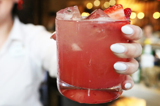 A waitress in a white shirt holds out a Red Cocktail with ice in a glass cup against a blurry background.