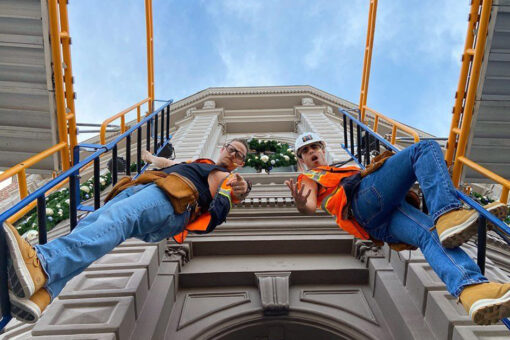 Two construction workers, both wearing jeans and yellow vests, climb ladders next to each other in front of a light-gray building. The worker on the building's right wears an orange belt of pouches, a black tank top, glasses, and has a mini microphone at his mouth. The worker on the building's left wears a white hard hat, and brown-ish-orange boots with a white bottom. There is a wreath in the center of the building's front window. There are various shapes adorning the fancy-looking building. The ladders on either side are blue, with black rungs, and lead to orange scaffolding. The sky is blue, with misty clouds.