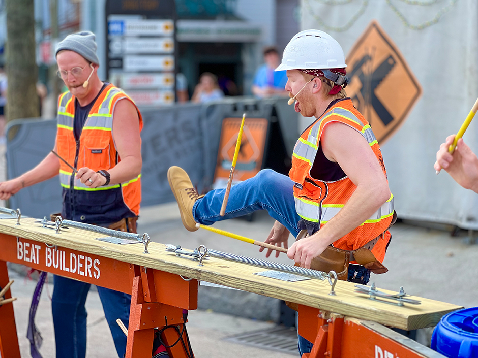 Two men in construction worker carb play the drums with light-brown-and-yellow drumsticks with white tips onto wood and metal construction materials. The man on the image's right wears a gray beanie, glasses, a microphone on his face, a watch, a silver ring, a dark blue shirt, an orange-yellow-and-silver construction vest, and jeans. The man on the left has his left leg, clad in blue jeans and a brown sneaker boot, in the air, while wearing a white hard hat, a red bandana, an earplug, a microphone on his face, a black tank top, and a matching vest to the first man. His drumstick is mid-toss, in the air, and his tongue is sticking out. He faces the first man. The hand of a third person, also holding a matching drumstick, is visible, though all else from that person is cut off. An orange, wooden sign reading "Beat Builders" in white, blocky text holds up the wood and tools being used as drums, and a second one is partially visible, along with part of an upside-down, blue garbage bin, but are also cut off. The background is blurred, though an orange diamond is visible on a canvas sheet. Within a black border almost on the edge of the diamond are black silhouettes of a hammer and saw making a shape in the letter "X," and two drumsticks crossed in another "X" shape in the center. Various pieces of blurry signage, as well as a tree, can be seen in the background too.
