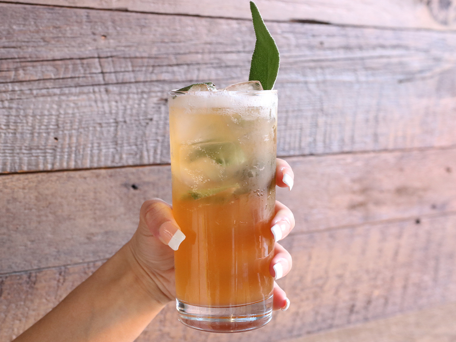 Photo of hand holding orange mocktail with ice and a leaf sticking out the top with wood wall in the background