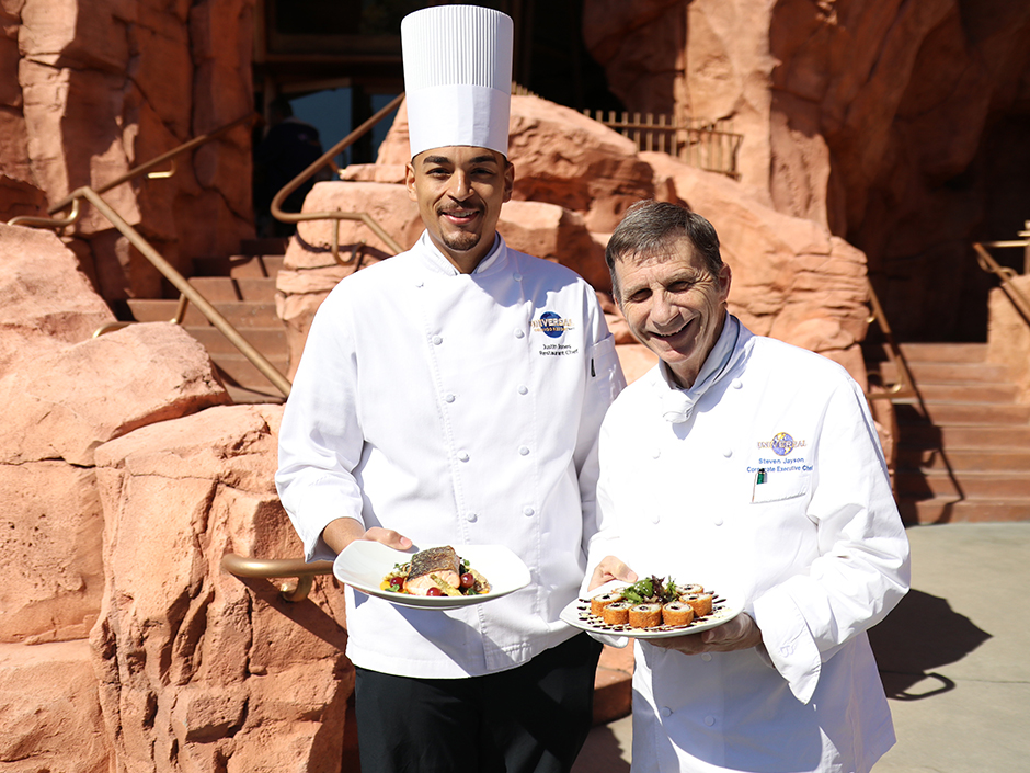 Chef Justin and Chef Steve holding plates with food in front of Mythos 