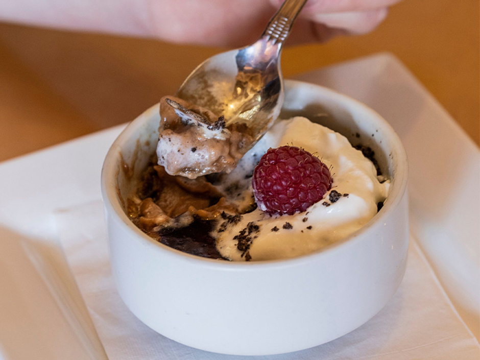 A silver spoon being lifted out of a creme brulee in a white bowl on top of a white napkin on a white plate, with some of the dessert sitting on the spoon. The dessert has a white cream, sprinkled chocolate, and a raspberry on top. The dish sits on top of a brown table.