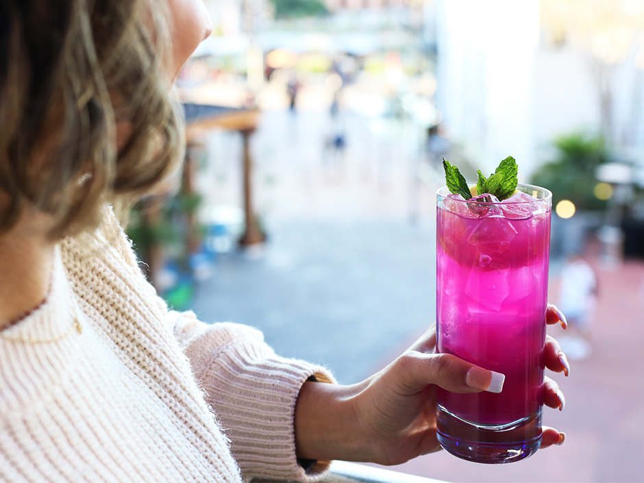 Photo of woman's profile holding pink drink with ice and leaves sticking out the top with CityWalk in the background. She wears a white sweater and necklace.