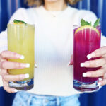 Photo of a woman in a white, long-sleeve shirt and jeans holding out a yellow drink and a pink drink against a blue background.