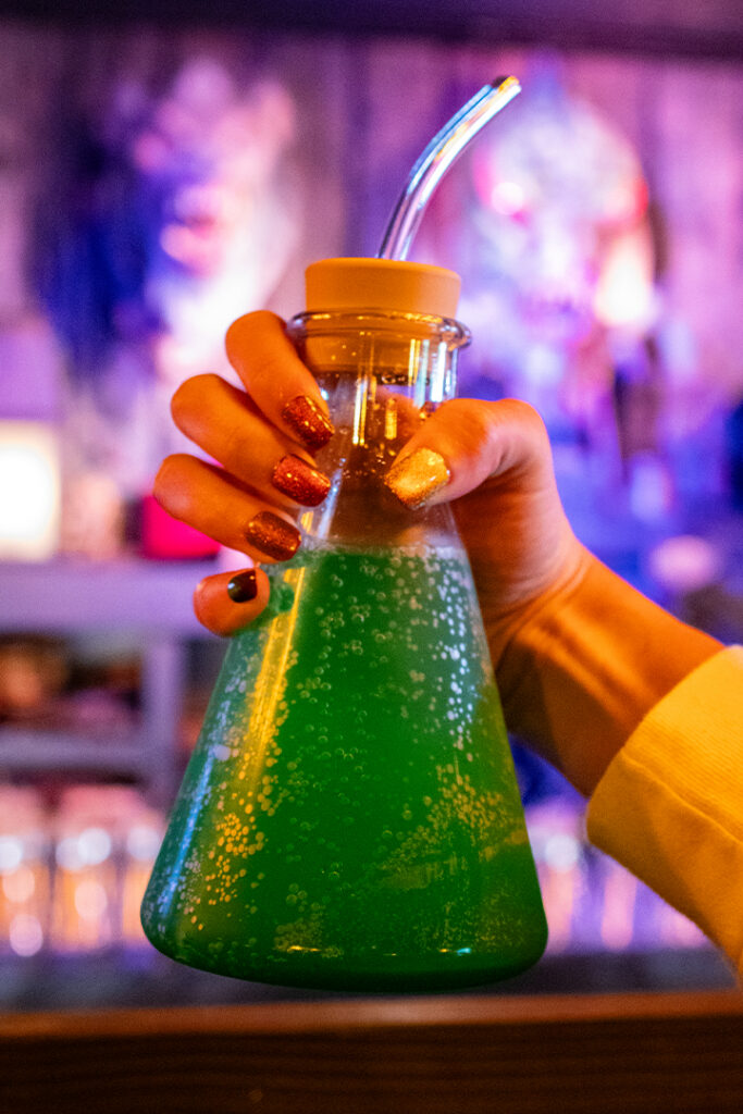 A hand holding up a Monocane Mocktail in a souvenir beaker glass inside Burning Blade Tavern