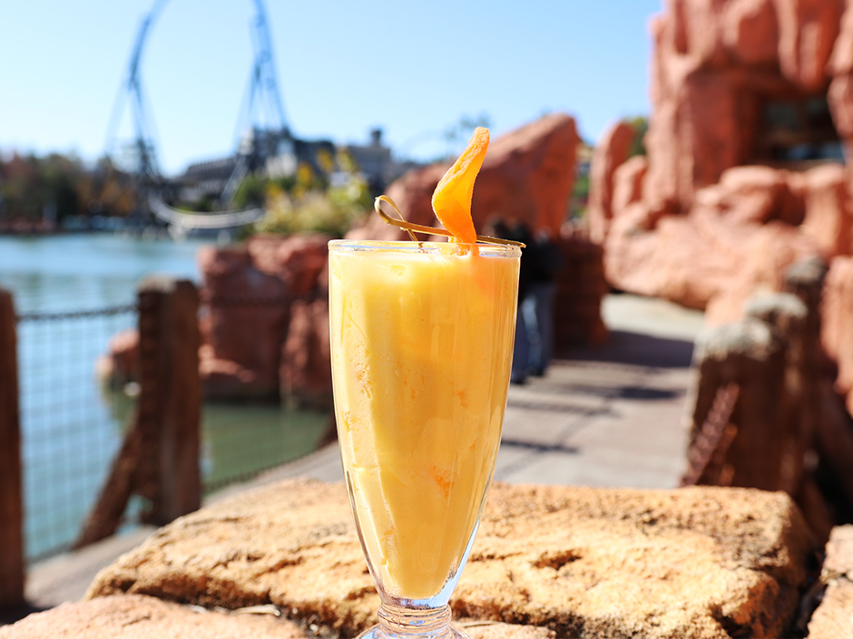 Orange mango lassi in glass outside Mythos Restaurant with a blurred Velocicoaster in the background. There is an orange object sitting on top of the glass, which stands on top of a light-brown, rock formation.
