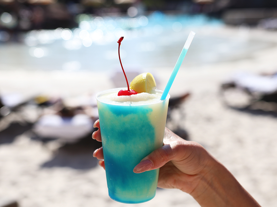 Hand holding frozen blue mocktail with Portofino's beach blurred in the background. There is a cherry on top of the drink, a straw in it, and a lemon on the cup's rim.