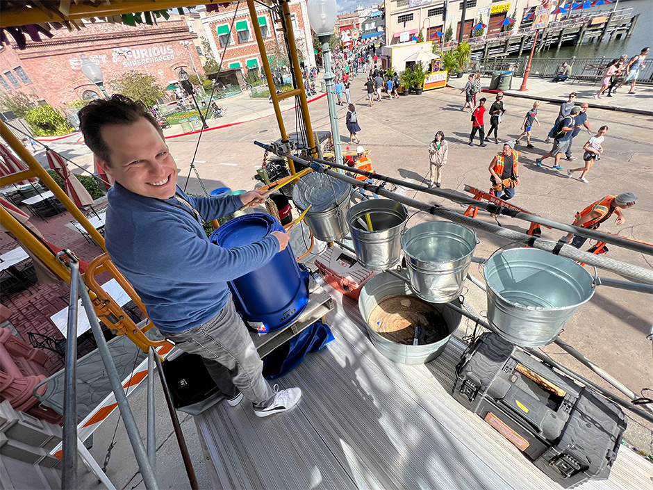 A man in a blue, long-sleeved shirt, gray pants ,and white sneakers with a black strip smiles at the camera, while playing the drums on a bucket with yellow drumsticks. The bucket is one of four that hang on a pole, high above the ground on scaffolding, in front of an upside down, blue garbage bin sitting on a wooden platform. Various toolboxes line the front of the scaffolding. The man's head is turned toward the camera, which is to his right, though his body faces forward, out to a group of people walking in front of a body of water and other buildings. Behind him, on the ground, are several seemingly marble tables with closed-up, red and beige table umbrellas and black chairs. To the tables' left is the red-brick building for Fast & Furious — Supercharged. A bit of a blue sky with clouds is visible far behind the buildings.