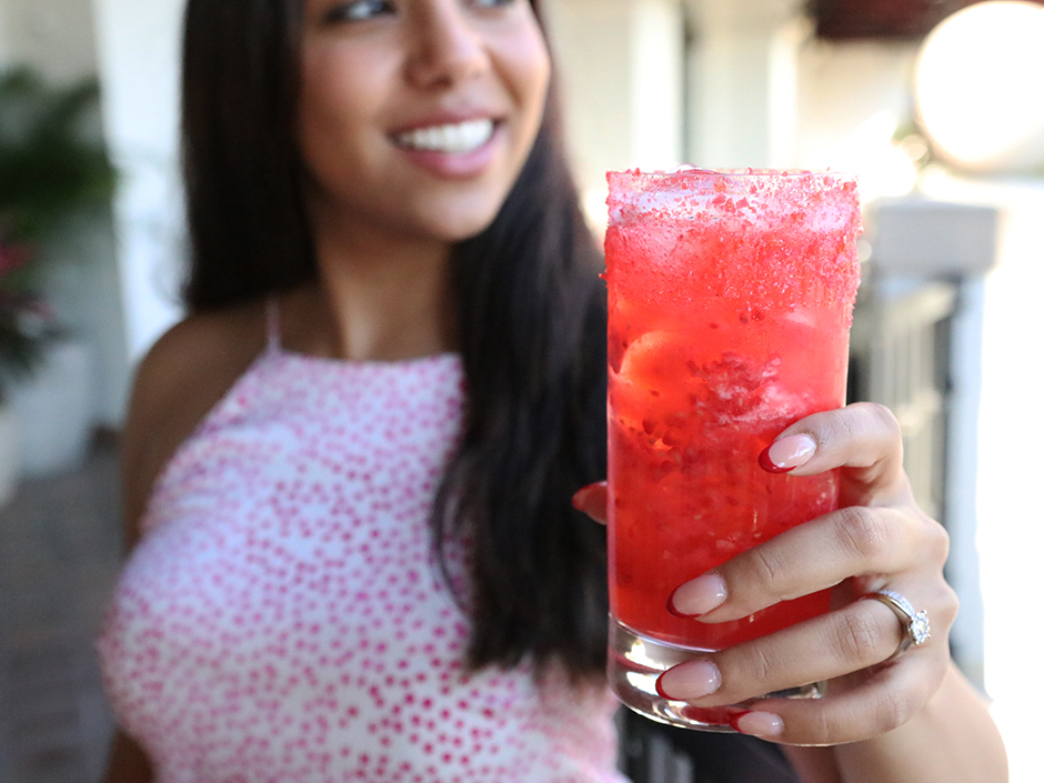 Woman in a white-and-pinkdotted dress holding red drink. The background is blurred, and the woman looks to her left, smiling.
