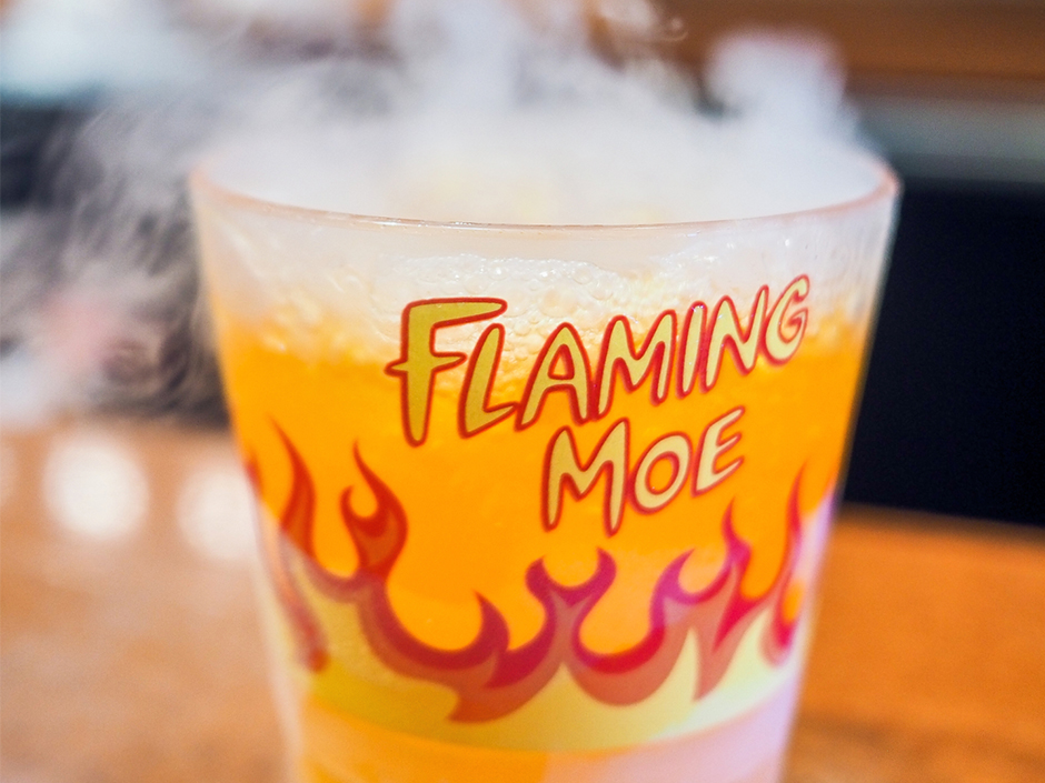Flaming Moe Orange drink with steam coming out of it. The cup has text "Flaming Moe" in yellow with a red background, and a fire image circles the glass.