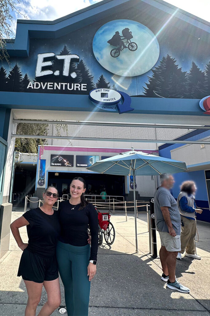 Two women stand slightly to the right of center, in front of the E.T. Adventure ride at Universal Studios Florida. The ride's sign is shaped like a house, painted with trees and a moon, in the center of which is a silhouette of a person riding a bike with something in the basket. Io its right is white, chalky text reading "E.T." and white blocky text under it reading "adventure." There is a circular wait time sign indicating 25 minutes, with the words "wait time" in blue, "0 2 5" on a screen, and "minutes" in the same blue. A blue arrow on its left points toward text reading "entrance" in dark blue over a light blue circle. Under the sign are two, horizontal metal poles. Under that is a light blue umbrella, with a silver pole. To its right is a folded up, but still standing, red-and-black wheelchair, and to its left is a black-and-white ride height indicator. Two blurred people stand in front of the height indicator, looking toward their left. Silver switchbacks lead to a large building entrance, on which is a pink border, and various E.T. signage. The woman on the right holds her right hand in a fist on her hip, and wears sunglasses, a short-sleeve black shirt, and black shorts. The woman on the left is younger, and wears a black, long-sleeve shirt, blue leggings, a necklace, and  a white smart watch.