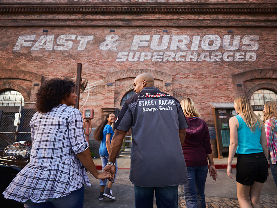 A group of people walk toward a brown, brick building with white, seemingly painted-on text reading "Fast & Furious Supercharged." The building has several half-circle, tile, window-like features. In front of the building are various gates, and signs indicating the current time and current wait time. Closest to the camera is a woman in a flannel-like patterned top and dark bottoms, holding hands with a man in a gray-and-black shirt that reads "street racing" in a blocky font and "garage service" in a curly font, along with other, harder-to-read text. Closer to the building, a younger woman with glasses, a blue shirt, jeans, and black sneakers, looks back toward a group of three women, all facing aware from the camera. From left to right: a woman in what looks like a purple sweatshirt and jeans, a woman in a teal tank top and black shorts, and a woman in a pink-white-and-turquoise flannel, and dark pants. There is what seems to be the top and front of a car to the woman closest to the camera's left. The floor is concrete, with some being seemingly made of a cobblestone. Right in front of the building is a lighter sidewalk, with a red curb.