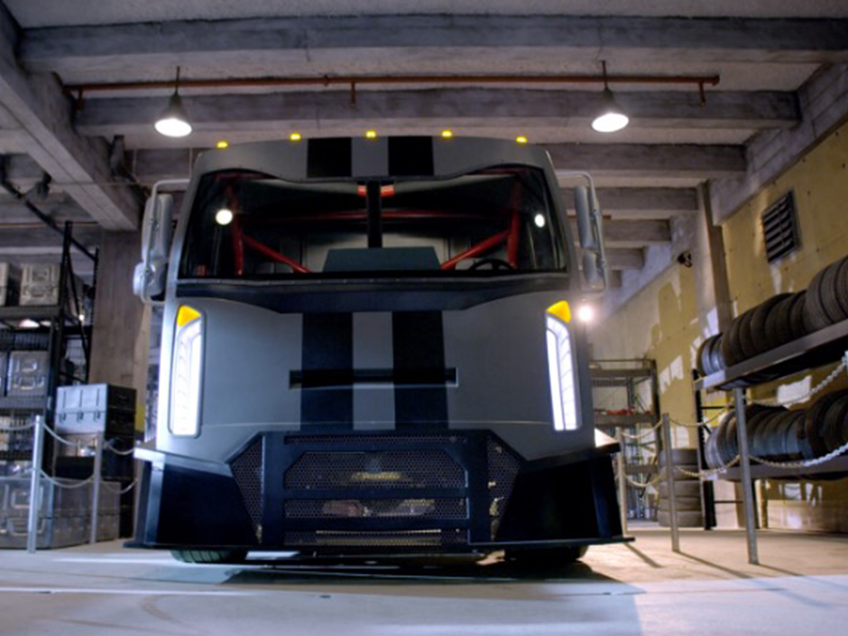 The front of the Fast & Furious — Supercharged Party Bus. The headlights are on. The bus itself is light gray, with two black, vertical stripes down the center, cut off by the glass windshield. There is no one visible in the driver's seat. The bus is inside of a garage, lit up by ceiling lights. There are shelves and stacks of tires to the bus' left, and cases to its right. Both sections are roped off. The ceiling and floor are both seemingly made of concrete, with the left wall being partially painted yellow.