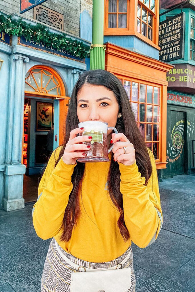 A woman in a yellow shirt and black-white-and-yellow patterned bottoms drinks butterbeer out of a clear mug. She stands in front of bright, multi-colored buildings at Diagon Alley in Universal Studios Florida.