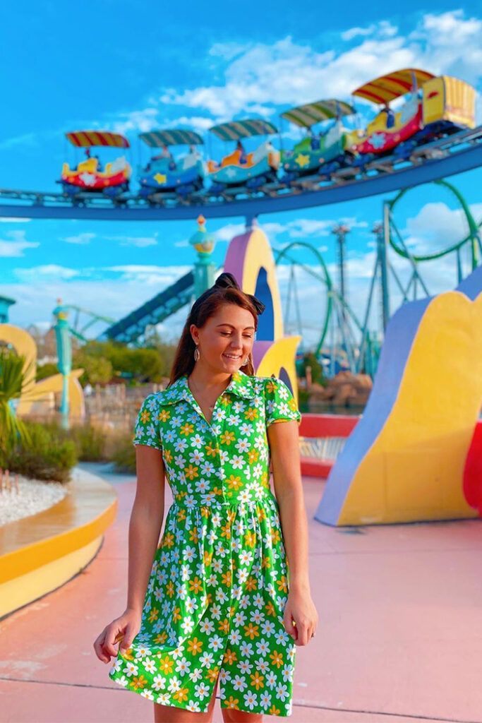 A woman wearing a black hair bow, circular earrings, and a green, button-up dress covered in orange and white flowers looks down and to her left, while pinching the bottom-right of her dress. She stands in Seuss Landing at Universal Islands of Adventure, surrounded by brightly colored structures and blurry plants in pebble patches. Behind her, the multi-colored and striped trolley vehicle of The High in the Sky Seuss Trolley Train Ride! passes by, containing five cars of differently colored versions of the same striped and starred patterns. The Incredible Hulk Coaster is visible in the distance, though blurry. The sky is bright blue, and has a few clouds.
