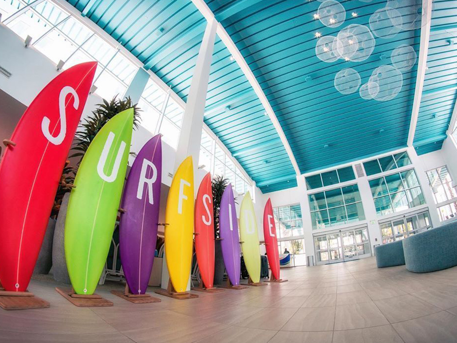 A view of a lobby at Universal’s Endless Summer Resort – Surfside Inn and Suites. Eight surfboards stand next to one another, each displaying a different white letter of the word "Surfside." From left to right, the surfboards are colored red, green, purple, yellow, red, purple, yellow, and red. The lobby has a white-and-turquoise color scheme, and the walls are covered in glass windows. Two turquoise, round ottomans sit on top of the light gray tiled floor. The ceiling is turquoise and paneled, and several almost-translucent, white circles can be seen in the near center of the ceiling. 