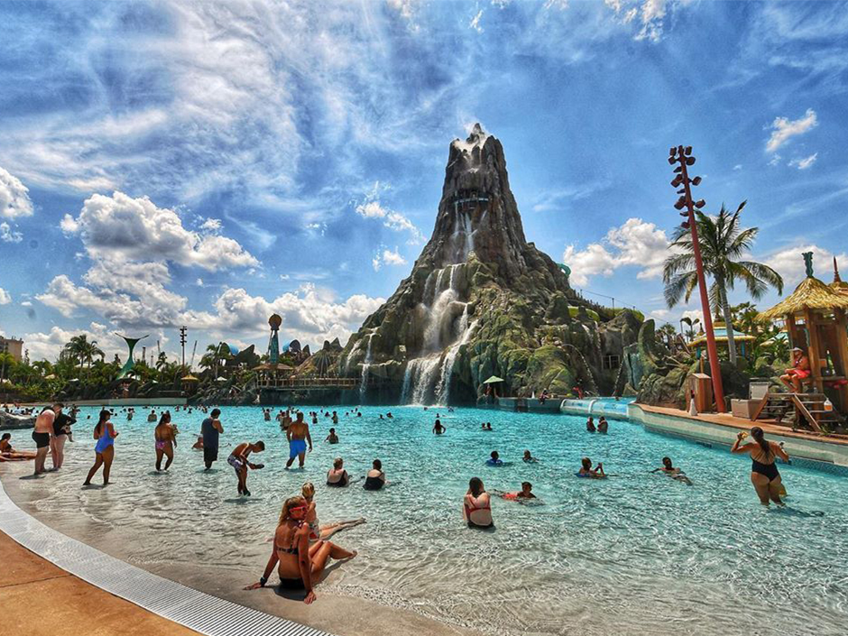A wide view of the Volcano and surrounding water at Universal Volcano Bay. Several people sit in and around the water. The sky is blue and full of white clouds. On either side of the volcano are several beach-style buildings and structures.