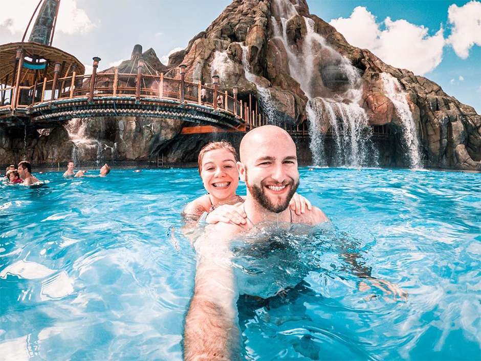 A man and woman stand smiling in a pool in front of the Universal Volcano Bay volcano, which has water falling down, seemingly from its unseen top.. They are both submerged up to their shoulders. The woman's hands are on the mans shoulders, and the man appears to be holding the camera to take their photo with him right arm. Other people stand and swim in the water, though separated from the pair. Behind the pair is a bridge, seemingly made of bamboo, decorated with various brown, white, and turquoise patterns. The right side of the bridge extends into a hole in the volcano, and people can be seen walking toward the hole. The left side of the bridge seems to end at a platform, which has a circular canopy, also seemingly made of bamboo. There is a hole at the canopy's center, with a turquoise strip lining it. A long tree-like structure sticks up from the top of the canopy. The sky is blue with white clouds.