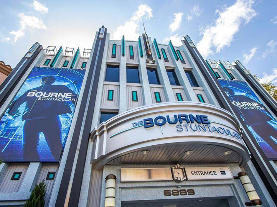 The Bourne Stuntacular building's entrance on a relatively clear day. The building has three connected towers, with the two side towers displaying signs reading "The Bourne Stuntacular" in white in front of a man in a jacket's back. On the signs, the man stands against a blue, gridded background, and has a circular target surrounding and slightly covering him. The silver-colored marquee on the center building reads "The Bourne Stuntacular" with the first and last words in light blue, and the middle word in darker blue and larger font. Under the marquee is an empty sign on the left, a clock in the center, and a sign on the right that reads "Entrance." Under that is brown text reading "6 8 8 9." Surrounding the sign and numbers are two cylindrical lamps. Above the marque are two rows of seemingly five, rectangular windows each, though only one window (and the corner of a second) are visible from the lower row. On all three buildings are thin, turquoise panels with dark borders. A stick with a pointed top sits at the center and top of the center tower.