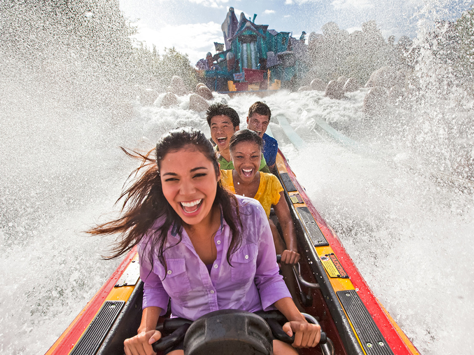 A red-and-yellow log-style boat full of guests after going down the hill on Dudley Do-Right's Ripsaw Falls. There is a massive splash of water on either side of the vehicle. From front to back in the log, there is a woman in a purple button-up shirt, a woman in a yellow shirt, a man in a green polo, and a man in a blue shirt. All four have their mouths open, smiling. Behind them is the hill they just came down, surrounded by the attraction's building. There are two waterfalls, surrounded by a cartoon-style house, with various signs, and mountain-like structures. The sky is blue, with a few clouds.