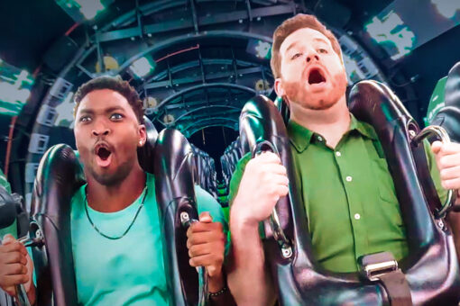 Two men have their mouths open, appearing to be screaming, while riding The Incredible Hulk Coaster at Universal Islands of Adventure. The man on the left wears a turquoise shirt and a necklace, and has an earring in his visible left ear. The man on the right wears a green button-up shirt with white buttons. Both men hold onto the handles on their thick, black shoulder restraints. They are riding through a tunnel with a techno visual theme and strips of what appear to be green lights. Rows of seats are visible behind them.