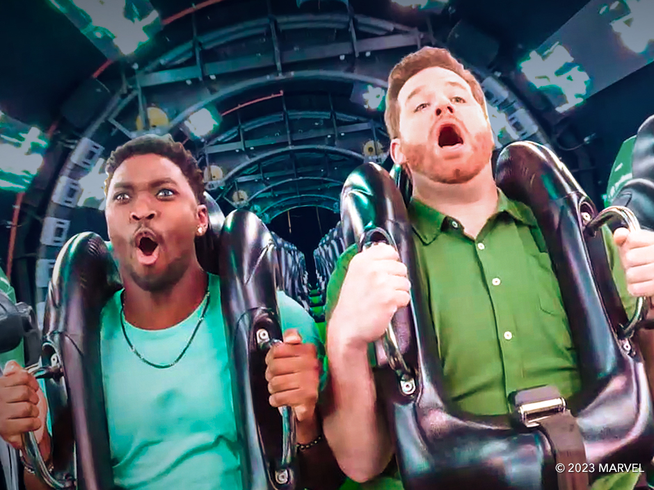 Two men have their mouths open, appearing to be screaming, while riding The Incredible Hulk Coaster at Universal Islands of Adventure. The man on the left wears a turquoise shirt and a necklace, and has an earring in his visible left ear. The man on the right wears a green button-up shirt with white buttons. Both men hold onto the handles on their thick, black shoulder restraints. They are riding through a tunnel with a techno visual theme and strips of what appear to be green lights. Rows of seats are visible behind them.