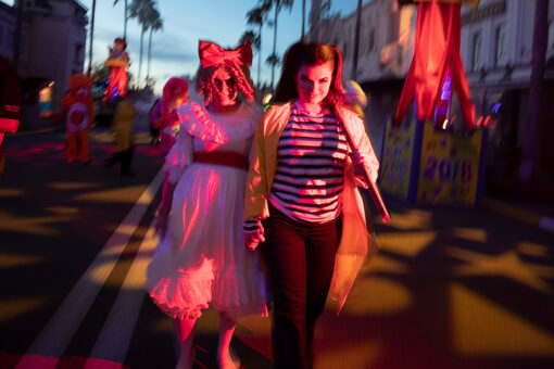 Two women walk down a street at Universal Orlando Resort during Halloween Horror Nights. The woman on the left wears what appears to be a light pink, ruffled dress with a red sash, and a bow in her curly, blonde hair. The woman on the left has her hair in half-up-half-down pigtails, wearing a pink shirt with red, horizontal stripes, leggings, and a yellow jacket. She is holding a mallet in her left hand. Behind the women are several creepy characters, some appearing like stuffed dolls come to life. In the background are several palm trees, and a fading blue sky with some gray clouds. Shadows line the street ground, on both sides of the double-white line running down the center. The surrounding builidngs are decorated with various Halloween-Horror-Nights-themed posters and decorations.