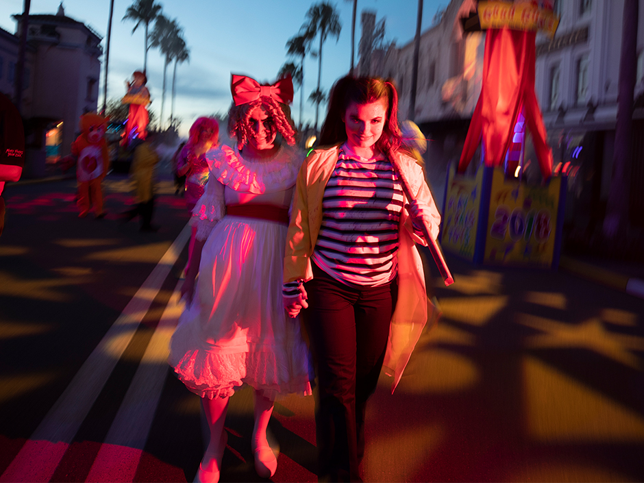 Two women walk down a street at Universal Orlando Resort during Halloween Horror Nights. The woman on the left wears what appears to be a light pink, ruffled dress with a red sash, and a bow in her curly, blonde hair. The woman on the left has her hair in half-up-half-down pigtails, wearing a pink shirt with red, horizontal stripes, leggings, and a yellow jacket. She is holding a mallet in her left hand. Behind the women are several creepy characters, some appearing like stuffed dolls come to life. In the background are several palm trees, and a fading blue sky with some gray clouds. Shadows line the street ground, on both sides of the double-white line running down the center. The surrounding builidngs are decorated with various Halloween-Horror-Nights-themed posters and decorations.