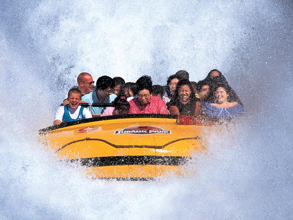 A yellow raft from Jurassic River Adventure full of guests, surrounded by a massive splash of water. The raft has black lining, a red number nine with a white border, and text "Jurassic Park" in white against a black rectangle with a white border, which sits on top of a red circle with a white border and a black dinosaur silhouette in the center.