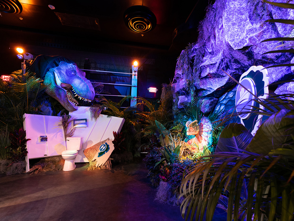 A view of a recreation of a "Jurassic Park" set at the Jurassic Park Tribute Store in Universal Studios Florida. The room is dark, with mostly purple lighting illuminating the set pieces and props. On the far left, in between plants and a gated railing with lights on either side, is the giant head of a dinosaur. It sits right above a set of a destroyed bathroom stall, with a closed toilet, surrounded by broken walls with a toilet paper dispenser, a seat cover dispenser, and a bathroom sign with the silhouette of a man and a woman. To the bathroom set's left, to the further left of more trees, is a rock formation. A smaller dinosaur head pops out of the bottom center of the rock structure, next to more plants on its other side. Above the dinosaur head and to its left is a plastic, two-layered sign. The bottom layer is navy, with a white border. The top layer is a lighter blue, also with a white border, with a white silhouette of a boat in the center.