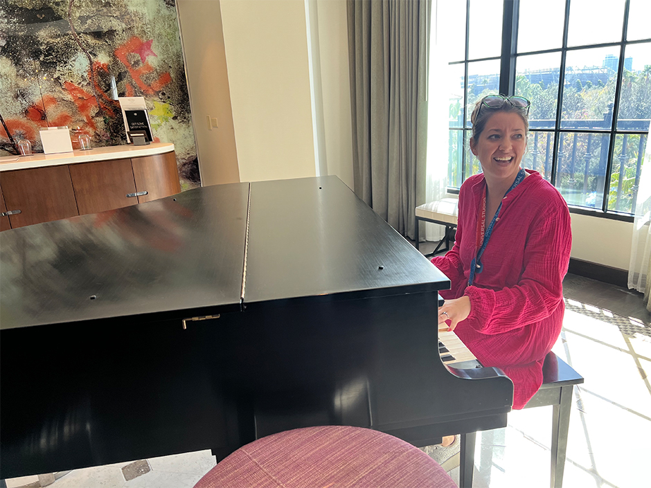 A woman in a pink, long-sleeved dress with glasses on the top of her head wearing a blue-and-red lanyard sits at a piano, with her hands on the keys. She looks her left, smiling. She, and the big black piano, are in a room with an open window looking out onto a gate and trees in front of a sunny sky, with solid curtains to the left and translucent curtains to the right. A small bench sits next to the solid curtains. On the back wall is the word "PEACE" in orange graffiti-style on a seemingly paint-splattered wall. In front of the wall is a round counter, with various books and trinkets. At the front and center of the photo is half of what appears to be a dark pink, round, cushioned stool.
