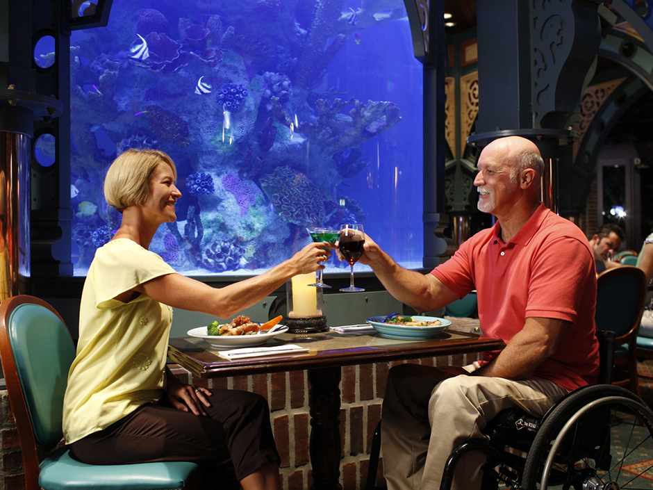A woman (on the left) and a man (on the right) sitting across from one another in front of a fish tank at Lombard's. The woman, who has short blond hair, is wearing a pale, yellow shirt with seemingly beaded flowers, and brown pants. The man, who has a light gray mustache and beard, is wearing a coral polo shirt, with khakis, and sits in what appears to be a wheelchair. The pair clinks their glasses together across the brown table, the woman's cocktail glass having a green liquid, and the man's glass having a red-ish-purple one. On the table are two plates of food, napkins, silverware, and a candle. Other diners can be seen behind the man, though mostly cut off and out of frame.