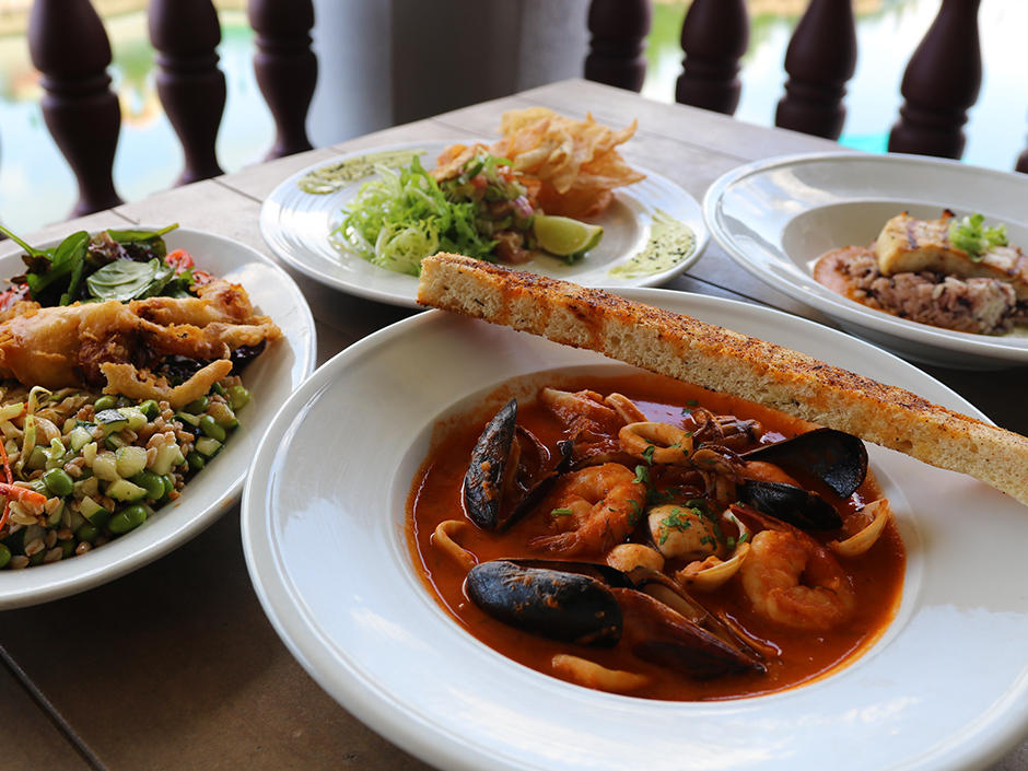 A view of four white dishes of food on a table. In the front and center is a bowl of primarily a red sauce, filled with various pieces of seafood. A piece of seasoned bread sits on top of the dish. on the left is a mix of various cut veggies, a fried crab, and a small side salad. In the back and center is a pile of greens, a piece of seafood that is tricky to decipher, and a handful of wonton chips. On the right is a piece of seasoned fish. Green onions sit on top of the fish, which itself sits on top of brown-and-black rice, within an orange sauce. Behind the table is a wooden deck railing. The rest of the background is blurred.