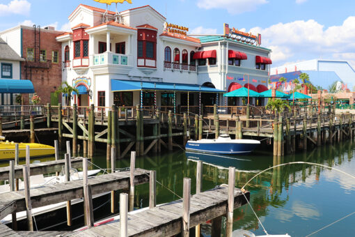 A wide view of Lombard's Seafood Grille at Universal Studios Florida. In front of the white building with turquoise and red awnings over the many windows, red lining, and a fish-shaped yellow sign with blue-ish-green text reading "Lombard's" on one of the roofs, is a body of water with a blue-and-white boat inside. Various wooden docs with wooden poles surround the water, and parts of boats floating on the water can be seen. The sky behind the building is blue, with a few white clouds.