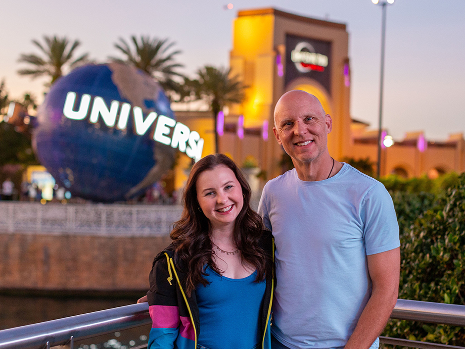 Meaghan Kelly, wearing a blue shirt, a necklace, and a blue, pink, and black Universal jacket, stands next to her father, who is wearing a light blue shirt on her left. They smile as they stand in front of a railing overlooking water at CityWalk. The father has his right hand on the railing. The Universal Globe and Universal Studios Florida Arch can be seen in the background, and the sky is fairly clear, though a bit dark.