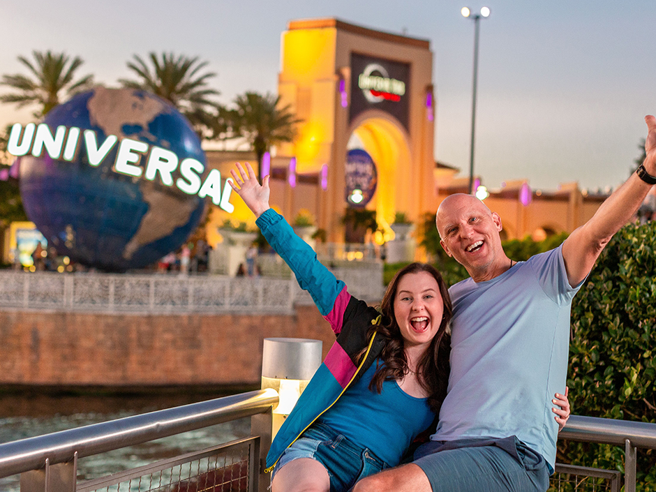 Meaghan Kelly, wearing a blue shirt, a necklace, jean shorts, and a blue, pink, and black Universal jacket, holds out her right arm in a diagonal above her head, and hugs her father with her left arm. Her father, wearing a light blue shirt and darker blue shorts, does the same thing in the opposite direction. They smile as they sit in front of a railing overlooking water at CityWalk. The Universal Globe and Universal Studios Florida Arch can be seen in the background, and the sky is fairly clear, though a bit dark.