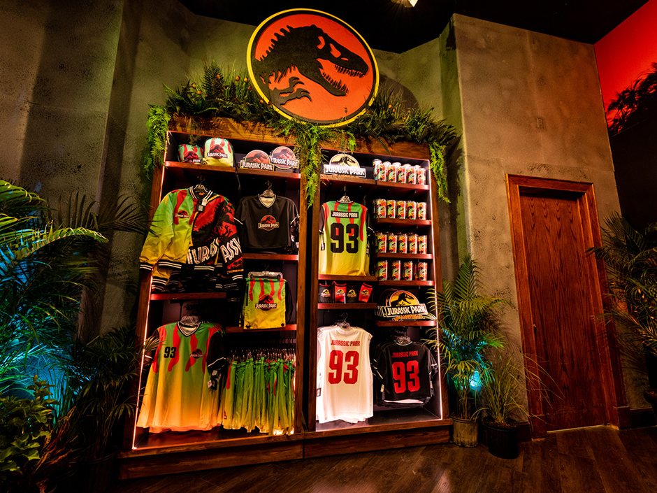 A view of a shelf displaying brightly colored "Jurassic Park" merchandise, standing on a wooden floor. Surrounding the shelf on all sides are various plants. On top of the shelf is an orange-ish-red circle with a black border surrounded by a yellow border, featuring a black silhouette of a dinosaur. The wall behind the shelf appears to be made out of a stone or rock, and there is a wooden door and doorframe to the shelf's left. On the far left, the beginnings of a background featuring red sky and a silhouette of plants can be seen.